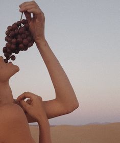 a woman holding grapes up to her face while standing in the desert with sand dunes behind her