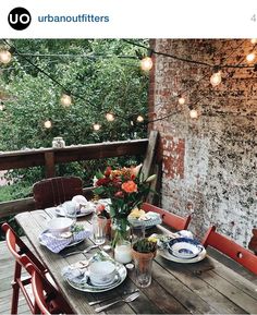 an outdoor table set for two with plates and flowers on it, surrounded by string lights