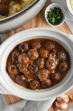 meatballs and gravy in a white bowl on a wooden cutting board next to other dishes