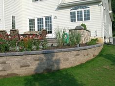 a large stone wall in front of a house with chairs and flowers on the lawn