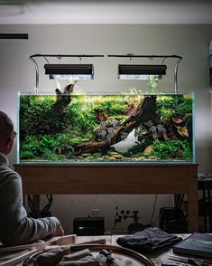 a man sitting in front of a fish tank filled with water and plants on top of a table