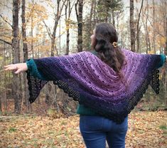 a woman is standing in the woods with her arms spread out, wearing a purple and green knitted shawl