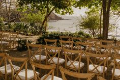 rows of wooden chairs with white cushions sit in front of an ocean and tree - lined area