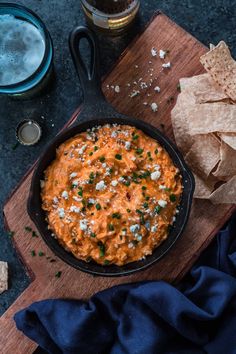 a skillet filled with dip and chips on top of a wooden cutting board next to a glass of beer