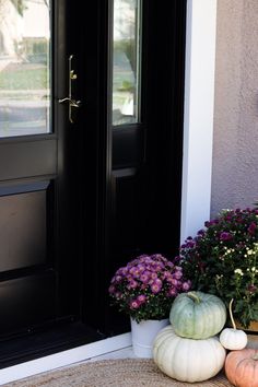 two white pumpkins and some purple flowers on the front step of a house with black doors