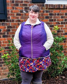 a woman standing in front of a brick building wearing a purple vest and floral skirt