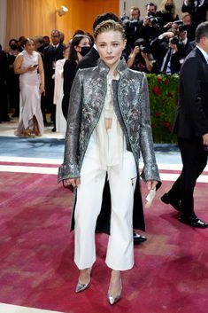 a woman standing on top of a red carpet wearing white pants and a silver jacket