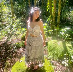 a woman in a white dress standing on mossy ground with trees and bushes behind her