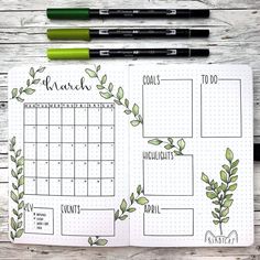 an open planner with green leaves on it and two markers next to it, sitting on top of a wooden table