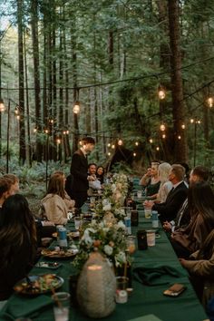 a group of people sitting around a long table in the woods with lights strung overhead