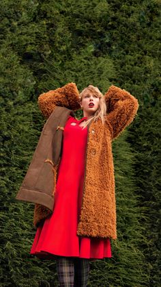 a woman in a red dress and brown coat is posing for the camera with her hands on her head