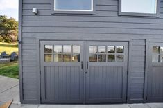 two garage doors on the side of a gray building with grass and trees in the background