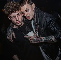 two men standing next to each other with tattoos on their arms and shoulder, both looking at the camera