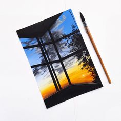 a painting with a pencil on top of it next to a window that looks out onto the sunset