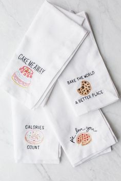 four embroidered tea towels on a marble countertop with cookies, cake, and donuts