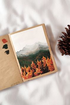 an open book sitting on top of a bed next to a pine cone and painting