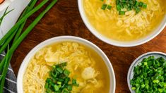 three bowls filled with soup on top of a wooden table next to green onions and carrots