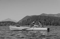 two people in a row boat on a lake with mountains in the background and trees