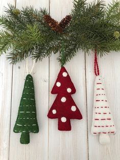 three felt christmas tree ornaments hanging from a pine branch on a white wooden table with pine cones