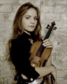 a woman with long hair holding a violin in front of a brick wall and looking at the camera