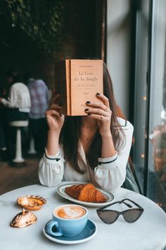 a woman sitting at a table with a book in front of her face and coffee