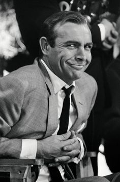 a black and white photo of a man in a suit smiling at the camera while sitting down