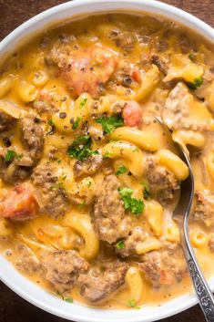 a white bowl filled with pasta and meat soup on top of a wooden table next to a spoon