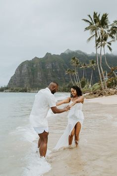 a man and woman holding hands in the water on a beach with mountains in the background