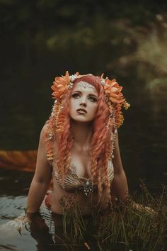 a woman with red hair and flowers on her head sitting in the water near grass