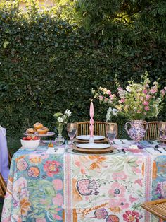 the table is set with plates, cups and bowls on it in front of a hedge