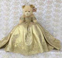 a teddy bear dressed in a gold dress sitting on a white wallpapered background