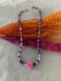 a beaded necklace with a pink smiley face on it and colorful feathers behind it