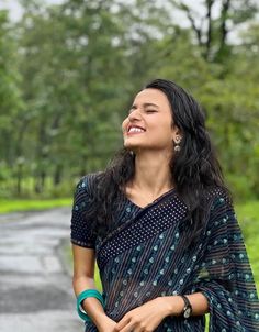 a woman standing on the side of a road smiling and looking into the distance with trees in the background