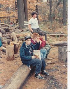 two boys sitting on a log in the woods with one boy holding a book and another boy standing behind them