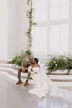 a bride and groom sitting on a bench in the middle of a room with large windows