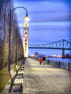 two people walking down a sidewalk next to benches and a clock tower in the background