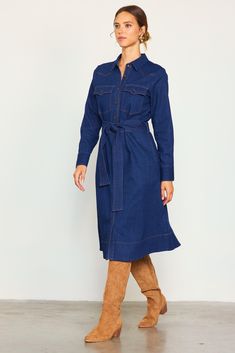 This denim shirt dress comes in a mid-length silhouette with long sleeves and a waist-cinching tie belt. Featuring a clean, dark wash, it showcases Western-inspired flap pockets and yoking accented by contrast topstitching. Button it all the way up to convey a refined look, or leave a few undone to keep things casual. •Pointed collar •Button front placket •Long sleeves with double-button cuffs •Angled yoke •Peaked flap pockets •Optional self-tie belt •Slight Stretch Item Number: 99975 Fabric Con Denim Blue Workwear Shirt Dress With Pockets, Denim Blue Shirt Dress With Pockets For Work, Workwear Denim Blue Shirt Dress With Pockets, Fall Denim Shirt Dress With Pockets, Knee-length Belted Denim Dress For Work, Medium Wash Shirt Dress With Pockets For Work, Belted Medium Wash Denim Dress For Work, Indigo Denim Dress For Workwear In Fall, Denim Blue Long Sleeve Shirt Dress For Work