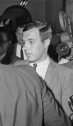 black and white photograph of man in suit sitting at desk with microphones behind him