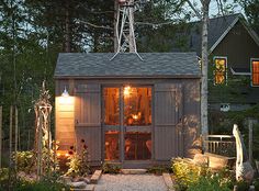 a garden shed with lights on the windows and plants in the yard next to it