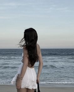 a woman walking on the beach carrying a black purse and looking at the ocean with her back to the camera