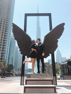 a man and woman standing next to each other in front of a statue with wings