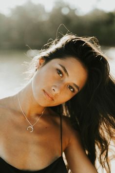 a woman with long dark hair wearing a black top and posing for a photo in front of the water