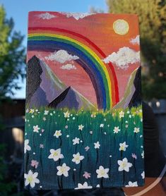 someone is holding up a painting with flowers in front of a rainbow and mountain range