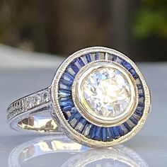 Details: Stunning vintage Art Deco style diamond & sapphire engagement ring. The band is platinum. This is an gorgeous ring with lovely engraving around the shoulders of the band, and has engraving details surrounding the diamond. This ring is stunning! Appraisal Reads: One old European cut diamond, measuring 7.20 x 7.20 x4.36mm an approx. weight 1.38ct. with faint blue fluorescences. Clarity H-I Cut Very Good Depth 60.6% Table 47.2% Six round brilliant cut diamonds measuring 1.5 x 1.5 x .90 Diamond Sapphire Engagement Ring, Blue Gemstone Rings, Engagement Ring Photos, Diamond Engagement Wedding Ring, Gorgeous Ring, Engagement Wedding Ring Sets, Sapphire Engagement Ring, White Gold Engagement, European Cut Diamonds