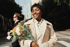 a man in a trench coat is holding flowers and walking down the street with another man behind him