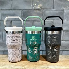 three travel mugs sitting next to each other on a wooden table with brick wall in the background