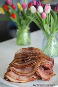 a white plate topped with sliced ham next to vases filled with tulips