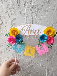 a hand holding a cake topper with paper flowers on it and the word ava spelled in large letters