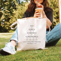 a woman sitting on the grass holding a tote bag and drinking from a straw