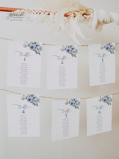 wedding seating cards hanging from a rope with flowers and leaves on the top, in front of a white wall